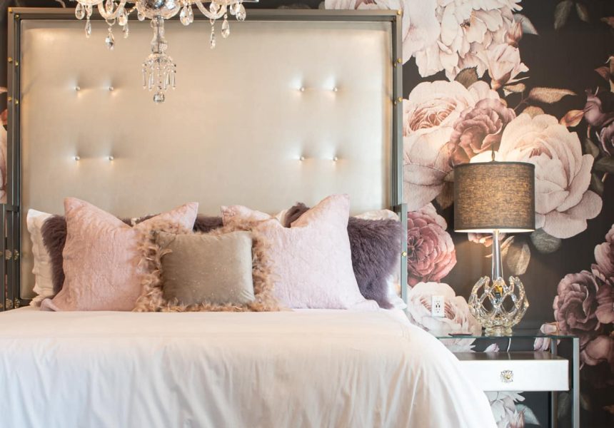 King bed with a plethora of pillows a chandelier and a glass end table set in front of floral decorative wallpaper