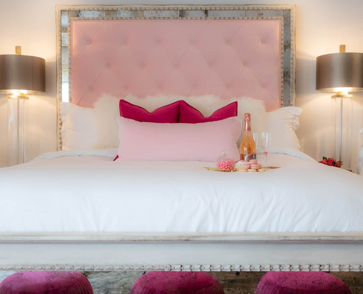 A king size bed with a pink fabric headboard two lamps on each end and 3 pink poofs at the foot of the bed