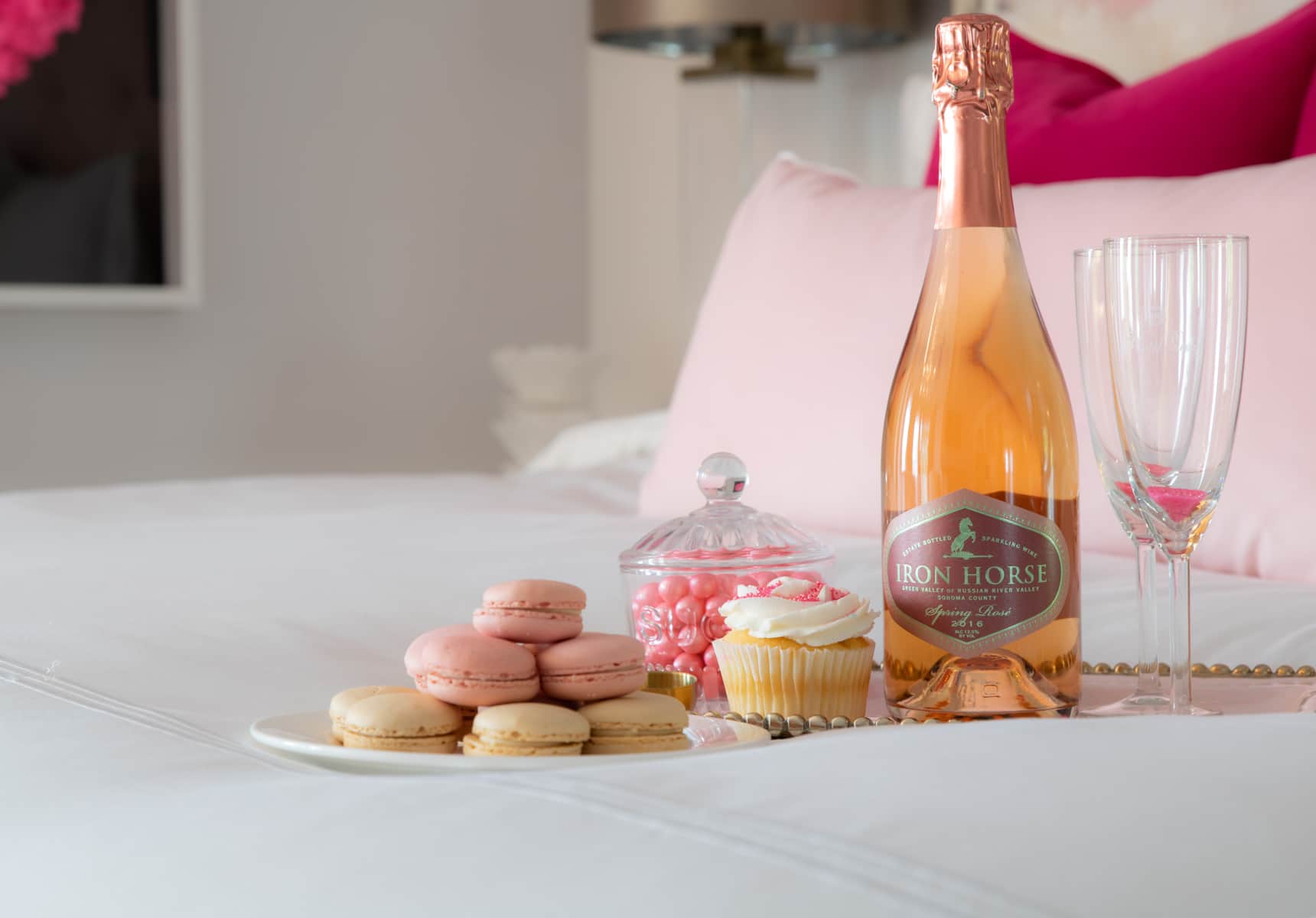 A small tray on a bed with french macaroons a cupcake two champagne flutes and a bottle of rosé